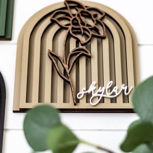 Op de afbeelding: Houten wandkunst met een gelaagd boogontwerp, een uitgesneden bloem en stengel, en de naam "Skylar" in cursief. Het kleurenpalet omvat beige, bruin en wit.