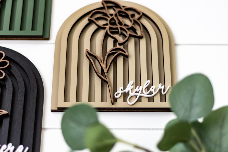 Op de afbeelding: Houten wandkunst met een gelaagd boogontwerp, een uitgesneden bloem en stengel, en de naam "Skylar" in cursief. Het kleurenpalet omvat beige, bruin en wit.