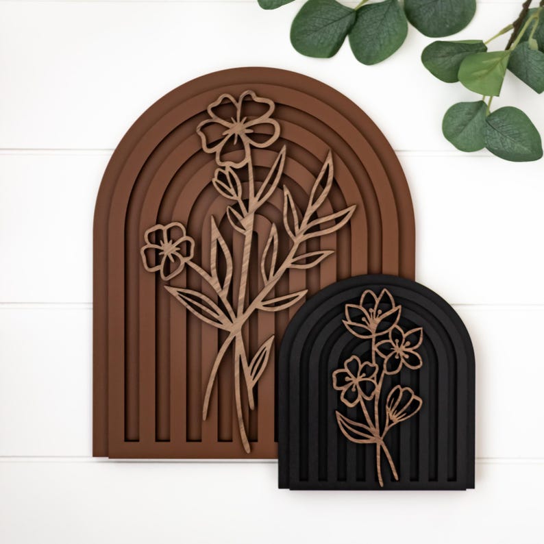 Op de afbeelding: Twee houten wandkunstwerken. E&eacute;n is een bruine boog met een bloemenontwerp, de andere is een zwarte boog met een bloemenontwerp. De bloemenontwerpen zijn uit licht hout gesneden. De achtergrond is wit.