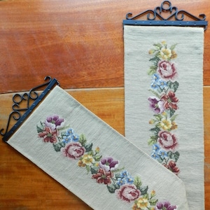 May include: Two beige needlepoint tapestries with floral designs, each hanging from a black metal rod.