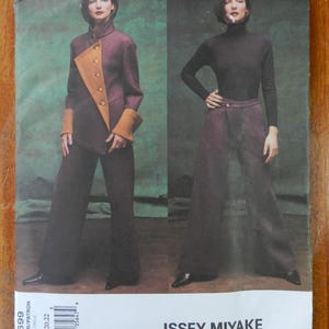 May include: A vintage sewing pattern featuring two women modeling Issey Miyake designs. One model wears a plum-colored jacket with tan accents and wide-leg pants. The other model wears a black turtleneck and matching pants. The pattern is labeled "ISSEY MIYAKE".