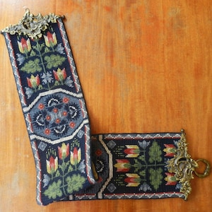 May include: A long, rectangular needlepoint bell pull featuring a dark blue background with repeating floral motifs in red, yellow, and green. The textile is finished with ornate, antique-style brass hardware at both ends, including a hanging ring.