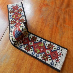 May include: A folded, rectangular textile with a detailed pattern. The design features red, green, and blue floral and geometric shapes on a beige background, framed by a black border. The textile appears to be hand-stitched.
