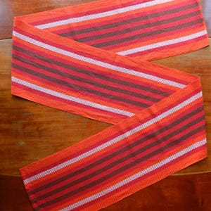 Könnte beinhalten: Ein langer, orangefarbener, rot-weiß-brauner gestreifter Tischläufer. Der Läufer ist aus einem gewebten Stoff gefertigt und hat eine strukturierte Oberfläche.