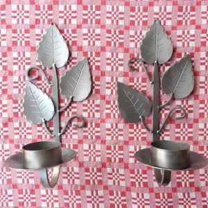 May include: Two silver metal candle holders with a leaf design. Each holder has a round candle cup and a decorative stem with three leaves.