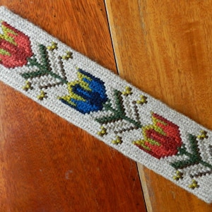 Könnte beinhalten: Ein beigefarbener Stoff-Lesezeichen mit gestickten Blumen in Rot, Blau, Gelb und Grün.