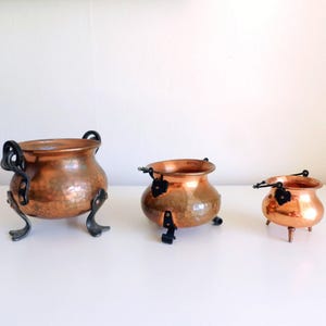 May include: Three graduated copper pots with a hammered finish and black metal accents. Each pot features a rounded body, a wide opening, and three sturdy legs. The two larger pots have decorative black handles attached to the sides.