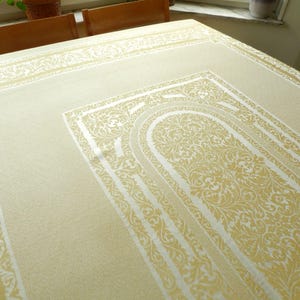 Puede incluir: Mantel crema con un diseño detallado y ornamentado en un tono crema más claro. El patrón incluye motivos florales y de volutas, enmarcados por bordes. El mantel está sobre una mesa de madera, con una ventana y plantas en el fondo.