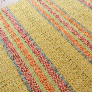 May include: A woven textile with a yellow background and vertical stripes in shades of pink, orange, green, and blue. The stripes have a textured, patterned design. The textile appears to be made of natural fibres.