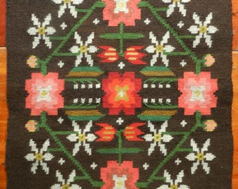 May include: A brown woven rug with a floral pattern. The rug features red, pink, white, and green flowers on a dark brown background.