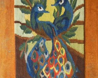 Op de afbeelding: Een geweven wandtapijt met twee blauwe pauwen op een tak. De pauwen hebben gedetailleerde staartveren in rood, geel en blauw. De achtergrond bevat een gele zon en groen gebladerte.