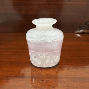 May include: A small glass vase with a rounded body and a flared rim. The glass features a swirling pattern of opaque white and translucent pale pink hues, accented by numerous small, clear air bubbles embedded throughout the material.