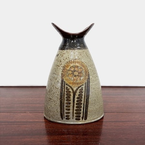 May include: A ceramic vase with a flared, dark brown rim and a speckled beige body. The front features a brown, stamped floral motif with a central circular flower and two vertical stems with leaf-like patterns extending downward to the base.