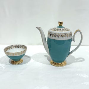 May include: A teal and white porcelain coffee pot with a matching small bowl. Both pieces feature a gold filigree pattern along the upper rim and a gold-toned base. The coffee pot has a tall, slender spout, a curved handle, and a lid with a gold knob.