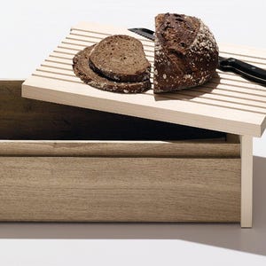 Può includere: Un porta pane in legno con un coperchio incernierato che ha un tagliere integrato. Il coperchio è aperto e una pagnotta di pane viene tagliata con un coltello.