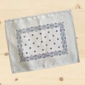 Può includere: Una tovaglietta rettangolare in tessuto beige chiaro con un bordo stampato blu scuro caratterizzato da intricati motivi a volute e puntini. Il centro della tovaglietta è decorato con un motivo ripetitivo di piccoli fiori a sei petali.