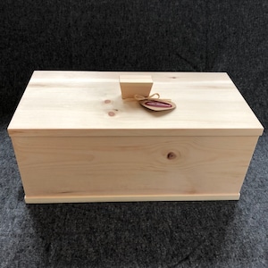 Puede incluir: Una caja de madera clara con una tapa con bisagras y un pequeño pomo de madera en la parte superior. La caja tiene una etiqueta en la parte delantera que dice "Holzwerkstatt".