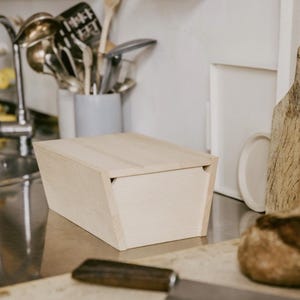 Könnte beinhalten: Eine helle Holzbrotbox mit einem Scharnierdeckel. Die Box ist aus Naturholz gefertigt und hat ein schlichtes, modernes Design.
