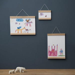 Op de afbeelding: Drie ingelijste kindertekeningen hangen aan een donkerblauwe muur. De tekeningen worden op hun plaats gehouden door houten magnetische posterhangers. De tekeningen bevatten kleurrijke illustraties van eenhoorns, een moskee en vlinders. Een ijsbeertje staat op de plank eronder.