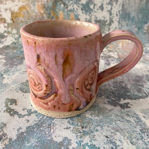 May include: A ceramic mug with a glossy pink glaze featuring two raised spiral relief patterns on the exterior. The mug has a curved handle and a speckled, unglazed base. The interior is smooth and finished in a matching light pink glaze.