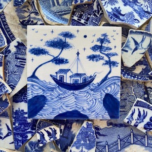 May include: Square ceramic tile featuring a blue and white illustration of a boat on wavy water, with trees and small houses. The tile is surrounded by broken pieces of blue and white ceramic. The design is reminiscent of traditional blue willow china.
