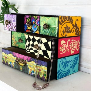 May include: A colorful wooden jewelry box with six drawers. Each drawer has a different patterned design, including floral, geometric, and paisley patterns. The box is decorated with gold-colored drawer pulls.