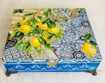 Puede incluir: Caja cuadrada de madera con tapa con bisagras, con un diseño de rama de limón. La tapa tiene un patrón de retazos con notas musicales, azulejos azules y una rama de limón con limones amarillos y hojas verdes. La caja es azul con adornos decorativos y patas metálicas.