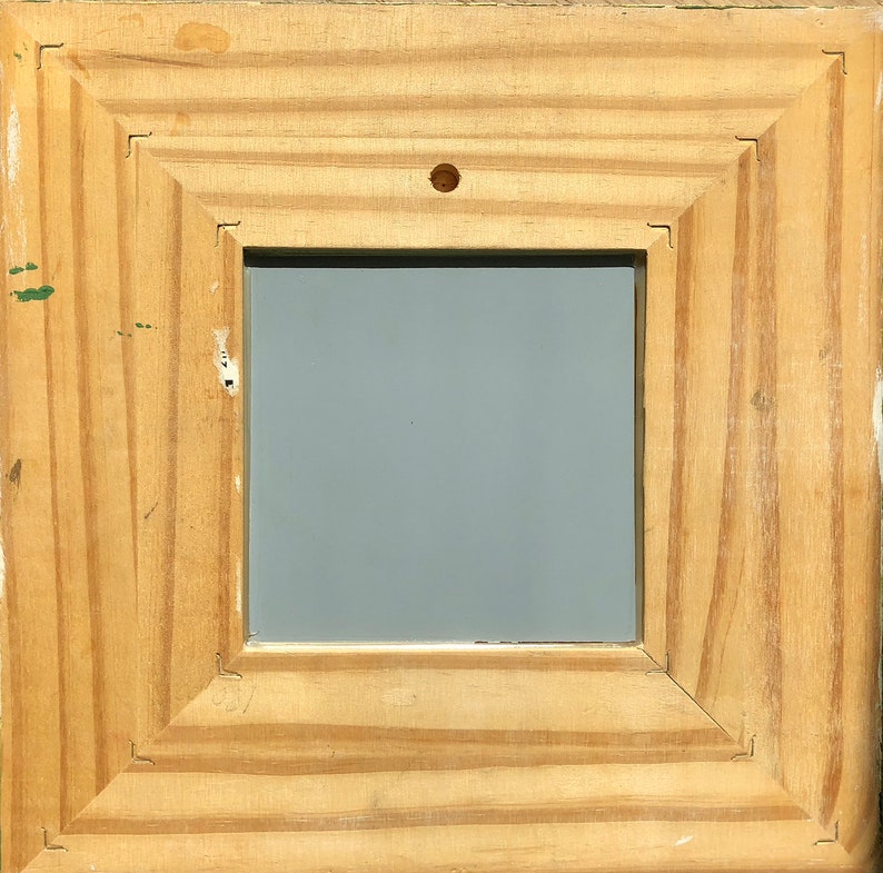 May include: Square wooden picture frame with a light brown finish. The frame has a layered design and a gray interior. A small nail is visible at the top center of the frame.