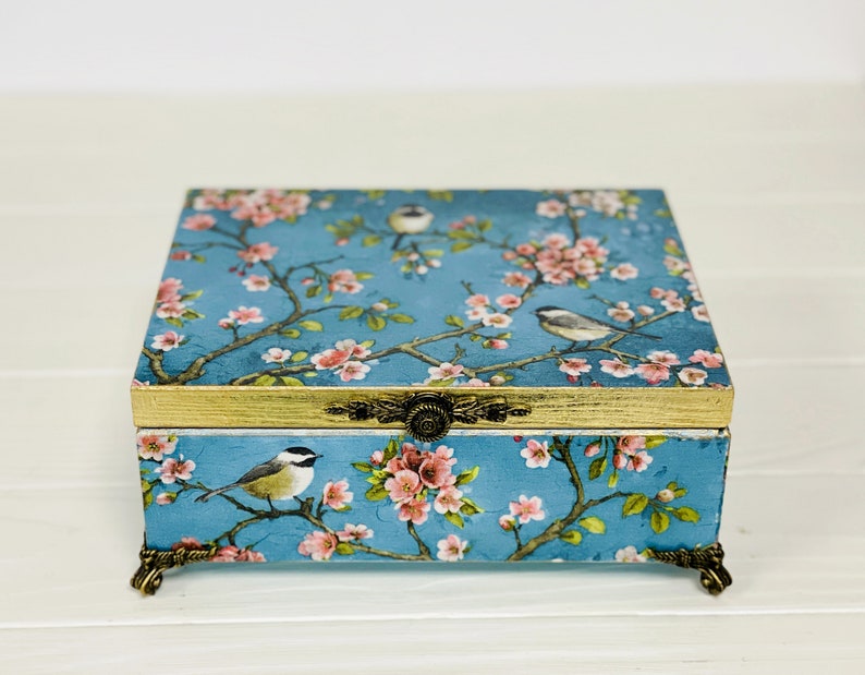 Puede incluir: Una caja rectangular con fondo azul y un patr&oacute;n de flores de cerezo rosas y p&aacute;jaros peque&ntilde;os. La caja tiene un borde pintado en dorado en la tapa, un cierre de metal dorado ornamentado y cuatro patas decorativas de metal dorado en la base.