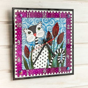 Könnte beinhalten: Quadratische Wanduhr mit einem skurrilen Katzendesign. Das Zifferblatt hat einen lila Rand mit weißen Punkten und einen schwarzen Rahmen. Die Katze ist schwarz und weiß mit grünen Augen, und der Hintergrund hat blaue Wirbel und rote Akzente.