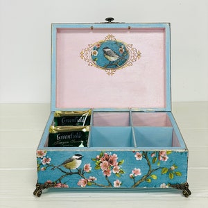 Puede incluir: Una caja de t&eacute; de madera azul claro con seis compartimentos, con un estampado floral y de p&aacute;jaros en el exterior y un interior rosa. El interior de la tapa muestra un p&aacute;jaro en un marco ovalado. Dos bolsitas de t&eacute; Greenfield est&aacute;n dentro.