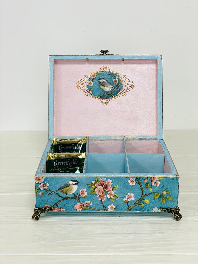 Puede incluir: Una caja de t&eacute; de madera azul claro con seis compartimentos, con un estampado floral y de p&aacute;jaros en el exterior y un interior rosa. El interior de la tapa muestra un p&aacute;jaro en un marco ovalado. Dos bolsitas de t&eacute; Greenfield est&aacute;n dentro.