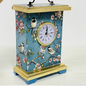 May include: A blue and gold wooden clock with a floral design and a bird motif. The clock has a handle on top and a quartz movement.