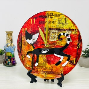 May include: A round clock with a black and white cat design. The clock face is red, yellow, and orange with black numbers. The cat is in a walking pose with a long tail. The clock is on a black stand.