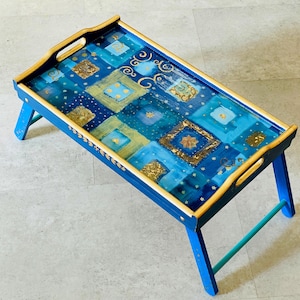 May include: A blue wooden tray with gold trim and a painted design of blue and gold squares. The tray has two foldable legs and a handle on each side.