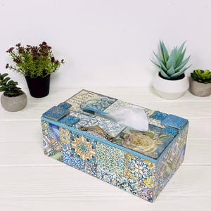 May include: A rectangular tissue box cover featuring a multi-colored mosaic tile pattern in shades of blue, yellow, and green, with a floral motif on the top, a metallic handle, and a central opening dispensing a single white tissue.