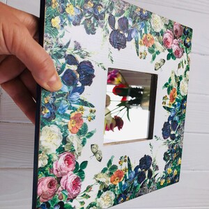 May include: Square decorative mirror with a floral design. The white frame is adorned with colorful flowers, including roses, irises, and pansies. Butterflies are also featured in the design. The mirror reflects a colorful bouquet.