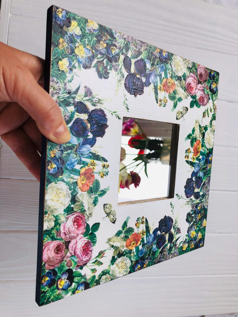 May include: Square decorative mirror with a floral design. The white frame is adorned with colorful flowers, including roses, irises, and pansies. Butterflies are also featured in the design. The mirror reflects a colorful bouquet.