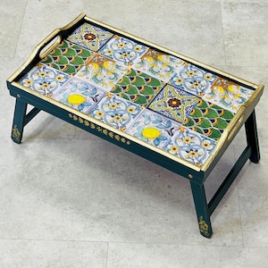 May include: A decorative wooden serving tray with folding legs. The tray features a colorful tile design with yellow lemons, blue flowers, and green foliage. The tray's frame is dark green with gold trim and measures approximately 61 cm long.