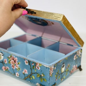 Puede incluir: Una caja de almacenamiento rectangular de madera con tapa abatible, con un exterior azul decorado con flores de cerezo rosas y p&aacute;jaros peque&ntilde;os. La caja tiene bordes dorados, patas de esquina de metal ornamentadas y un interior morado claro.