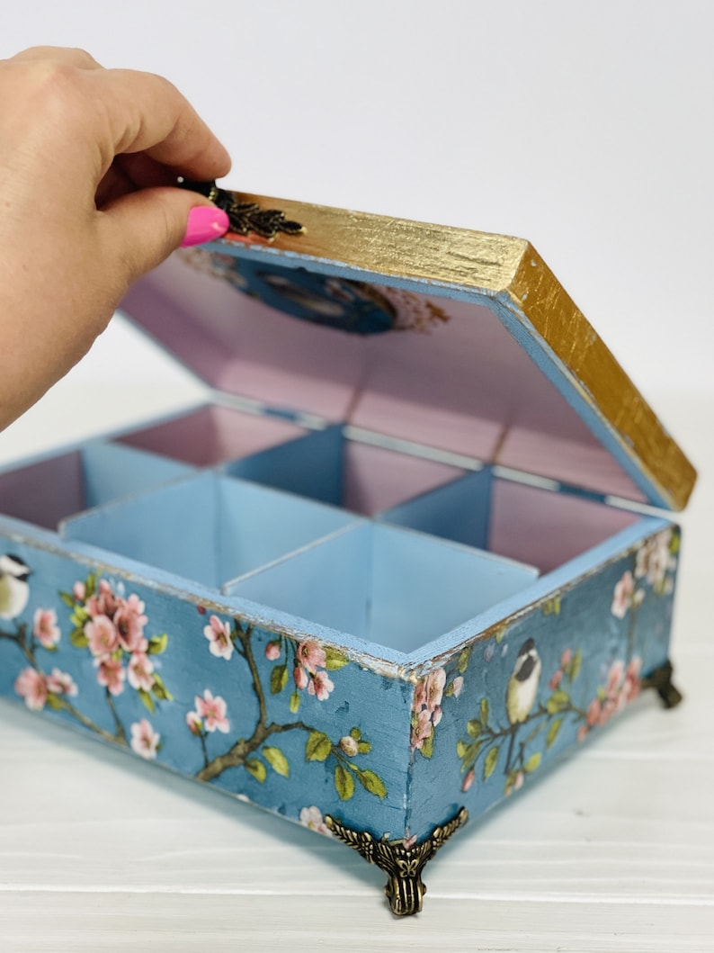 Puede incluir: Una caja de almacenamiento rectangular de madera con tapa abatible, con un exterior azul decorado con flores de cerezo rosas y p&aacute;jaros peque&ntilde;os. La caja tiene bordes dorados, patas de esquina de metal ornamentadas y un interior morado claro.