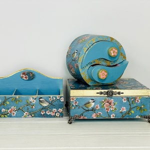 Puede incluir: Un conjunto de tres cajas de almacenamiento de madera azul con un patr&oacute;n de flores y p&aacute;jaros. El set incluye un organizador rectangular abierto, una caja rectangular con tapa y patas ornamentadas, y un cofre circular de dos cajones.
