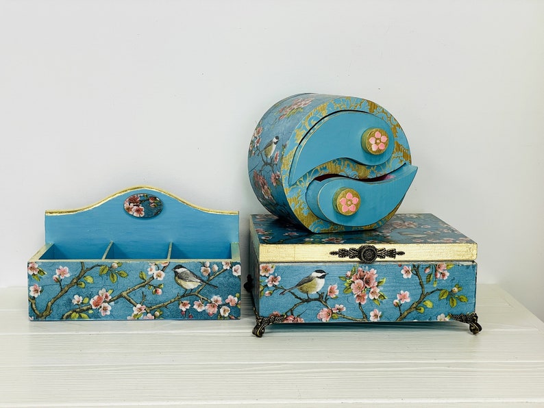 Puede incluir: Un conjunto de tres cajas de almacenamiento de madera azul con un patr&oacute;n de flores y p&aacute;jaros. El set incluye un organizador rectangular abierto, una caja rectangular con tapa y patas ornamentadas, y un cofre circular de dos cajones.