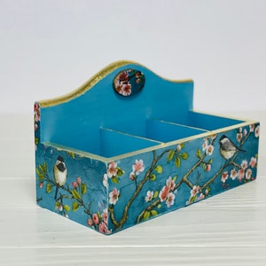 Puede incluir: Un organizador rectangular de madera con tres compartimentos, con un acabado azul y un patr&oacute;n floral de flores rosas y p&aacute;jaros peque&ntilde;os sobre ramas. El panel trasero es m&aacute;s alto con un borde superior curvo y un detalle ovalado.