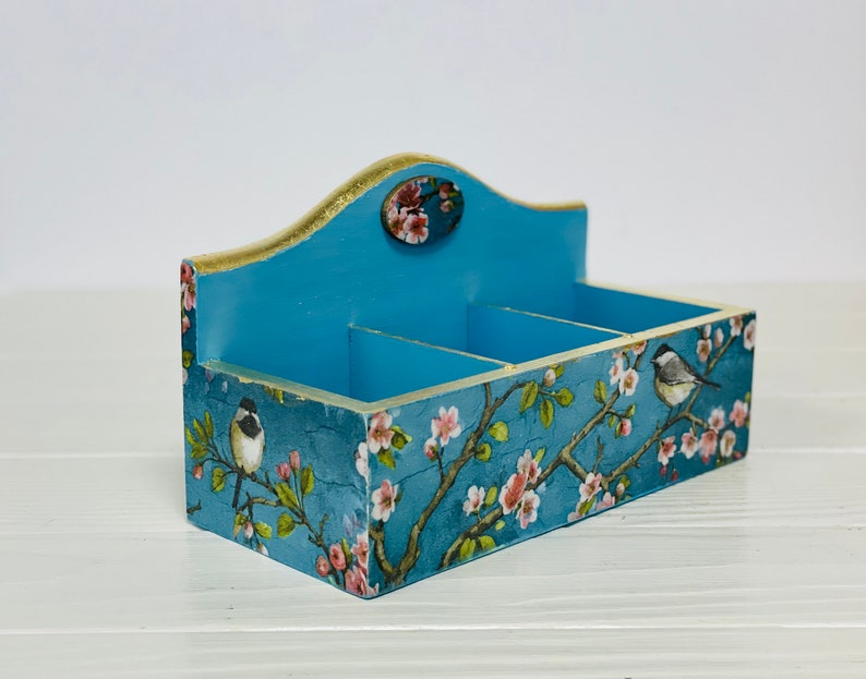 Puede incluir: Un organizador rectangular de madera con tres compartimentos, con un acabado azul y un patr&oacute;n floral de flores rosas y p&aacute;jaros peque&ntilde;os sobre ramas. El panel trasero es m&aacute;s alto con un borde superior curvo y un detalle ovalado.