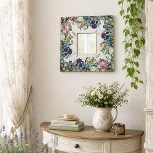 May include: Square wall mirror with a floral design frame featuring pink, purple, and blue flowers. The mirror hangs above a wooden table with a vase of white flowers, books, and a candle holder. The room has a vintage, cottagecore aesthetic.
