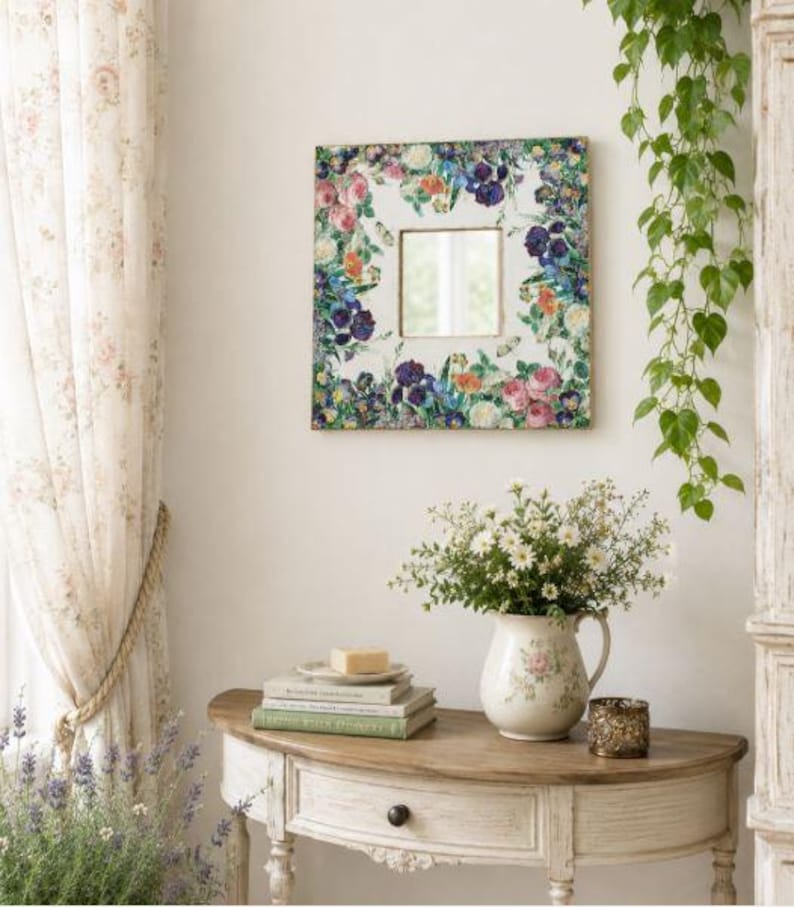 May include: Square wall mirror with a floral design frame featuring pink, purple, and blue flowers. The mirror hangs above a wooden table with a vase of white flowers, books, and a candle holder. The room has a vintage, cottagecore aesthetic.