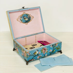Puede incluir: Una caja rectangular de madera con tapa abatible, pintada en un acabado azul desgastado con motivos florales y de aves. El interior est&aacute; forrado en rosa claro, con una ilustraci&oacute;n de un p&aacute;jaro en la tapa y patas decorativas de metal.