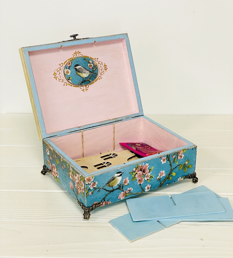 Puede incluir: Una caja rectangular de madera con tapa abatible, pintada en un acabado azul desgastado con motivos florales y de aves. El interior est&aacute; forrado en rosa claro, con una ilustraci&oacute;n de un p&aacute;jaro en la tapa y patas decorativas de metal.