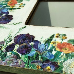 May include: A white wooden picture frame with a floral design. The frame is decorated with a collage of colorful flowers, including purple irises, pansies, and orange poppies. The frame has a distressed, vintage look.